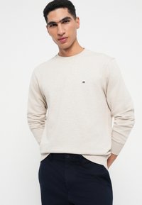 Felpa crewneck beige chiaro in misto cotone, con maniche lunghe e un piccolo logo sul lato sinistro del petto. Indossata con pantaloni blu navy.