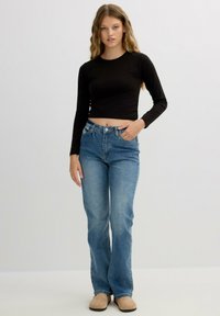 Top corto de manga larga negro combinado con jeans de campana de color azul claro y zapatos slip-on beige. La modelo se encuentra con una mano en la cadera.