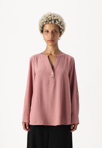 Langermet, rosa bluse med spiss utringning, to dekorative knapper, avslappet passform, og myk tekstur, sammen med mørke bukser.
