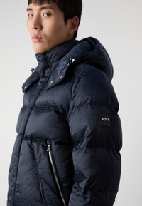 Mörkblå puffjacka med huva, med ett geometriskt mönster, blixtlåsförsedda sidofickor och en svart "BOSS" logotypbricka på ärmen.