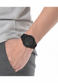Montre noire avec un bracelet en caoutchouc texturé et un cadran rond, présentant un logo vert et des marquages argentés sur un cadran noir.