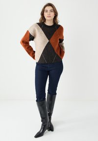 Pull en tricot avec un design en blocs de couleurs comprenant des losanges orange, beige, noir et marron. Col montant et coupe ample. Associé à un jean noir et des bottes noirs jusqu'au genou.