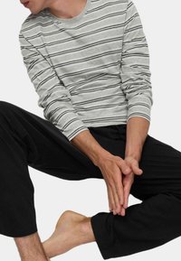 Camicetta grigia a righe lunghe con righe bianche e nere, abbinata a pantaloni neri larghi. Uomo seduto, con caviglia e mani in vista.