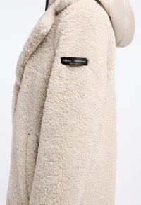 Beige schapenvachtjas met een zachte textuur, voorzien van een capuchon en een zwart label op de mouw met de tekst "FRIEDA FREDDIES NEW YORK."
