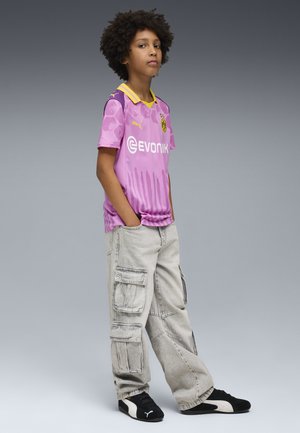 Lila Fußballtrikot mit gelben Akzenten und grafischem Design, kombiniert mit hellgrauen Cargo-Hosen mit mehreren Taschen und schwarzen Turnschuhen.