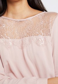 Top rosa pallido con un velo di pizzo trasparente decorato con motivi floreali e un tessuto liscio e leggero. Design a barca morbido con maniche lunghe.