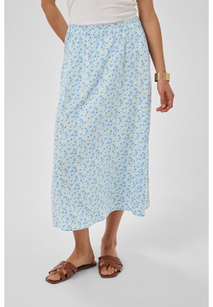 Frau trägt einen hellblauen floralen Midirock, ein weißes ärmelloses Oberteil, braune geflochtene Sandalen und ein breites goldenes Armband am rechten Handgelenk.