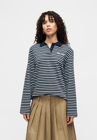 Vrouw met krullend haar, gekleed in een marineblauw-wit gestreept poloshirt met lange mouwen en een beige geplooide rok, staande tegen een effen achtergrond.