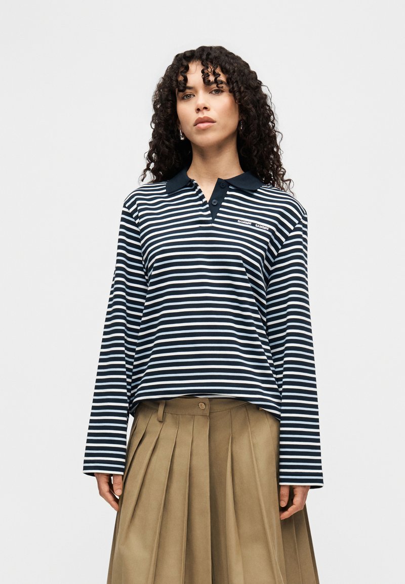 Vrouw met krullend haar, gekleed in een marineblauw-wit gestreept poloshirt met lange mouwen en een beige geplooide rok, staande tegen een effen achtergrond.