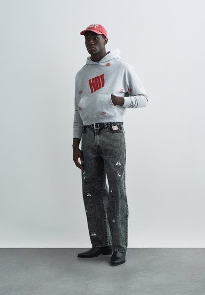 Grå cropped hettegenser med rød "HOT"-tekst og mønster av lepper, kombinert med svarte denimjeans med hvite motiver og et svart naglebånd. Sorte støvler.