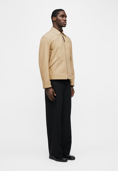 Άνδρας που στέκεται φορώντας μπεζ μπουφάν με φερμουάρ, μαύρο  παντελόνι με φαρδιά μπατζάκια και μαύρα παπούτσια, μπροστά σε ένα απλό λευκό φόντο.