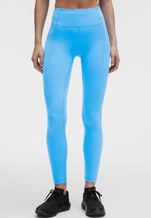 Kvinde iført lyseblå højtaljede leggings med sidelommer og sorte sportssko står mod en lys ensfarvet baggrund.