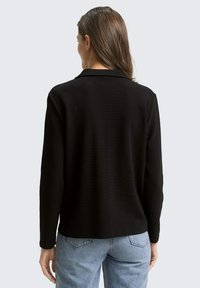 Schwarzes Langarmoberteil mit strukturierter Oberfläche, mit einem Kragen und geradem Schnitt, kombiniert mit hellblauen hoch taillierten Jeans.