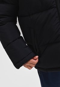 Melna pūkaina jaka ar teksturētu virsmu, sānos ar rāvējslēdzēja kabatu un aprocēm uz atsevišķām piedurknēm. Tuva rokas sasaite ar rāvējslēdzēju.