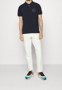 Marinblå polotröja med korta ärmar och en cirkulär logotyp; vita byxor; färgglada sneakers med gröna, röda och gula accenter.