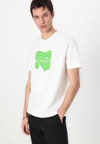 Bílý bavlněný tričko s velkým zeleným geometrickým logem a textem "KARL LAGERFELD JEANS" přes hrudník. Krátké rukávy, kulatý výstřih.