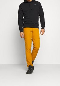 Haut à capuche noir avec un logo sur la poitrine, associé à un pantalon jaune slim avec des coutures au niveau des genoux. Des chaussures noires complètent la tenue.
