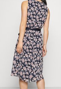 Květinové šaty s tmavě námořnickým pozadím, zdobené růžovými a bílými květy, bez rukávů a s černým páskem v pase. Měkká, splývavá tkanina.