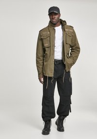 Olive green military-style jacket with multiple pockets, paired with black cargo pants and combat boots. Accessories include a black cap.