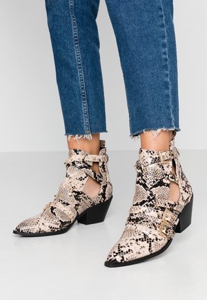 Jalat, joissa on kärjestä terävät, käärmekuvioiset nilkkurit, joissa on kultaiset soljet ja leikkausyksityiskohdat, yhdistettynä repaleisiin, sinisiin farkkuihin.