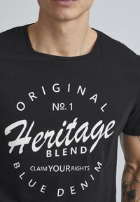 Crna pamučna majica s bijelim grafičkim natpisima: "Original No. 1 Heritage Blend Započnite svoja prava Plavi traper" u raznim fontovima.