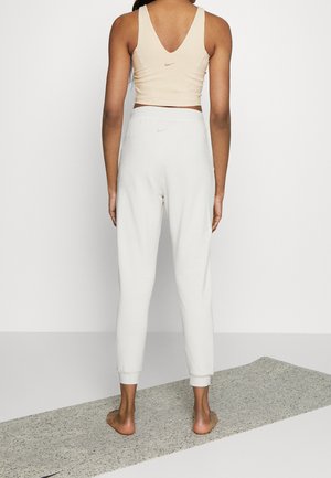 Femme debout pieds nus sur un tapis de yoga, portant un débardeur beige et un pantalon de survêtement blanc, tournée de dos à la caméra.