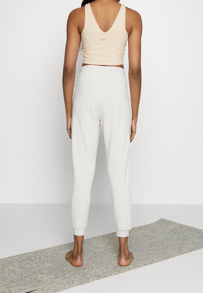Femme debout pieds nus sur un tapis de yoga, portant un débardeur beige et un pantalon de survêtement blanc, tournée de dos à la caméra.