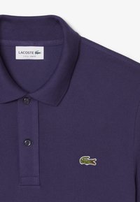 Lila bomullspolo med klassisk krage, två svarta knappar och en grön krokodil-logotyp på vänster bröst. Slim fit design.