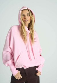 Růžová oversized mikina s kapsou vpředu, šňůrkami a žebrovanými manžetami. Má vyšívaný text v červené barvě. Doplněna tmavě hnědými kalhotami.