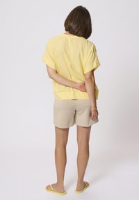 Haut jaune à manches courtes, ample ; shorts beiges ; tongs jaunes. Le modèle se tient les mains derrière le dos, tourné vers l'arrière. Design simple.