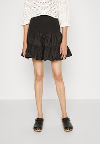 Vrouw in een zwarte gelaagde minirok, witte geborduurde cropped blouse en zwarte leren klompen met houten zolen tegen een effen achtergrond.