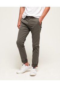 Chinos grigio scuro con vestibilità slim, orli arrotolati e dettagli rossi sulle tasche, abbinati a scarpe da ginnastica bianche. Tessuto di cotone morbido.