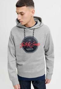 Siva dukserica s kapuljačom i vezicom, s kružnim crno-crvenim grafičkim logotipom sprijeda. Izgleda mekano i teksturirano.