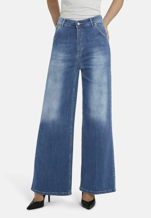 Jean bleu à jambes larges avec des panneaux avant délavés, porté avec des talons noirs à bouts pointus et un haut blanc, les mains dans les poches.