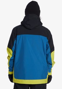 Blauwe, zwarte en neon gele jas met een capuchon; heeft een kleurenspelontwerp en elastische manchetten; achteraanzicht, textuurstof.