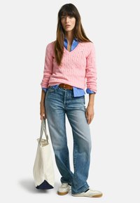 Růžový svetr s pleteným vzorem přes světle modrou košilí s límečkem, modré džíny a bílé tenisky. Drží krémovou tašku s námořnickým dnem.