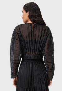Zwarte geplisseerde blouse van doorschijnende stof, met lange mouwen, elastische tailleband en een striksluiting bij de hals. Subtiele textuur met een gelaagde uitstraling.