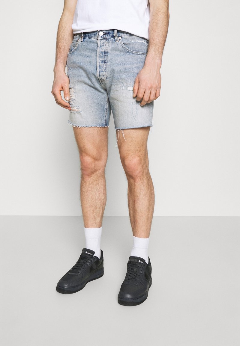 Shorts en denim de couleur bleu clair avec une finition déchirée et un ourlet effiloché ; associés à des baskets noires et des chaussettes blanches à la cheville.