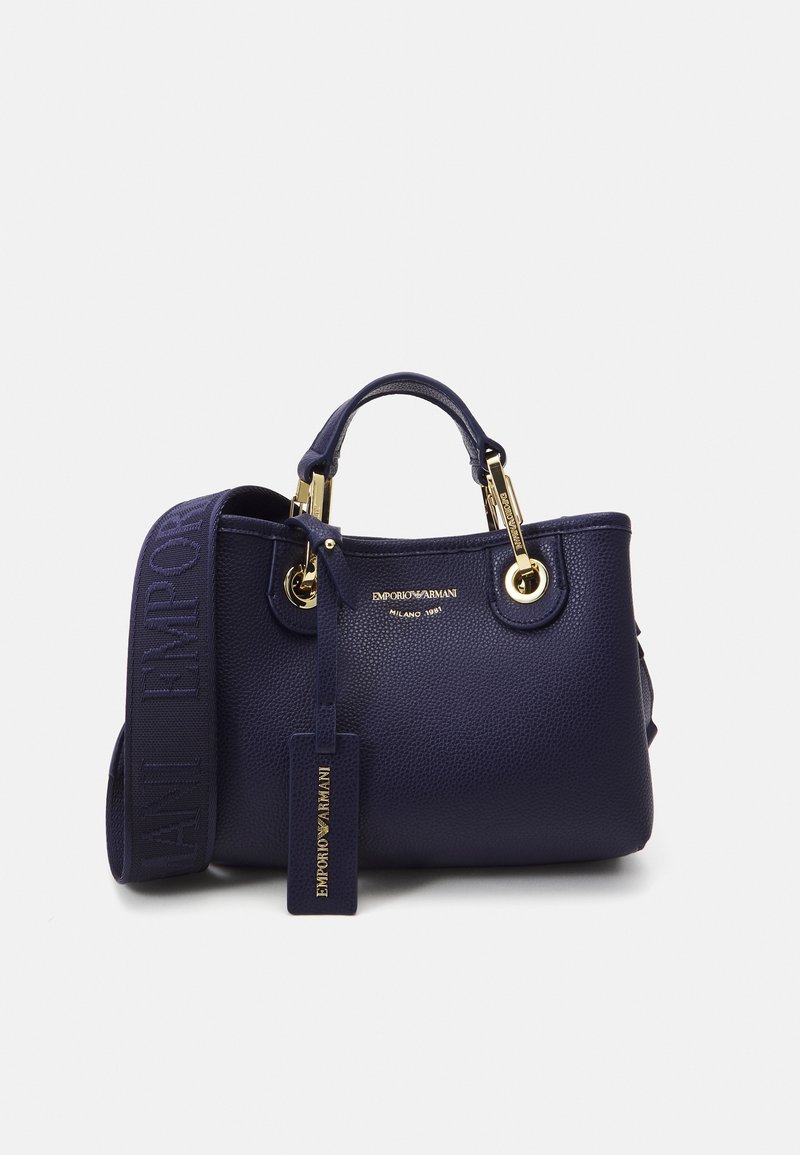 Námořnická modrá kabelka z texturované kůže se zlatým hardwarem, dvojitými horními rukojeťmi a nastavitelným ramenním popruhem. Obsahuje embosovaný štítek s logem.