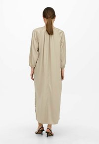 Lange beige jurk van een gladde stof, met pofmouwen, een geplooide halslijn en zijsplitten. Gecombineerd met zwarte sandalen met hak.