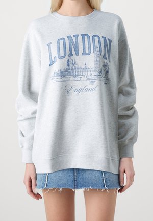 Jasnoszara bluza z niebieskim tekstem "LONDON England" oraz grafiką panoramy Londynu, noszona z postrzępioną jeansową mini spódniczką na osobie.