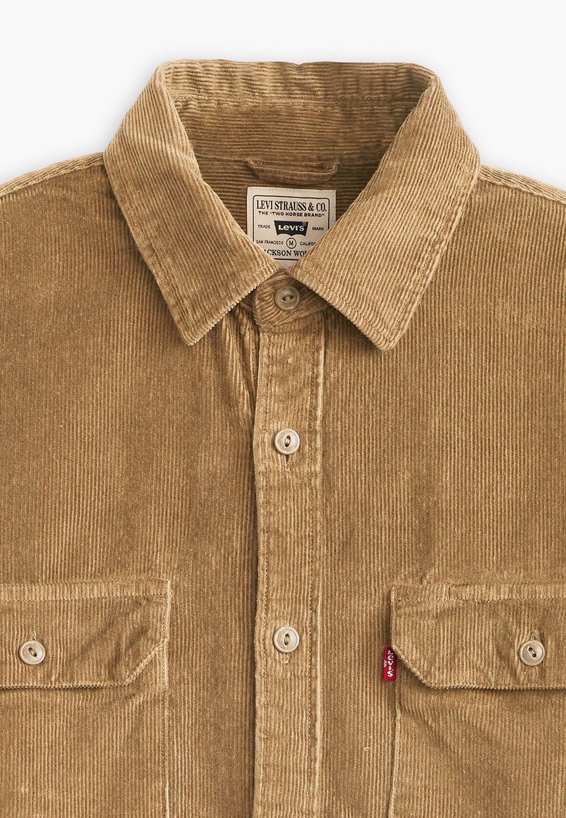 Camisa de pana marrón con cuello, cierre frontal de botones y dos bolsillos en el pecho con solapas de botón. Pequeña etiqueta roja de Levi's en el lateral.
