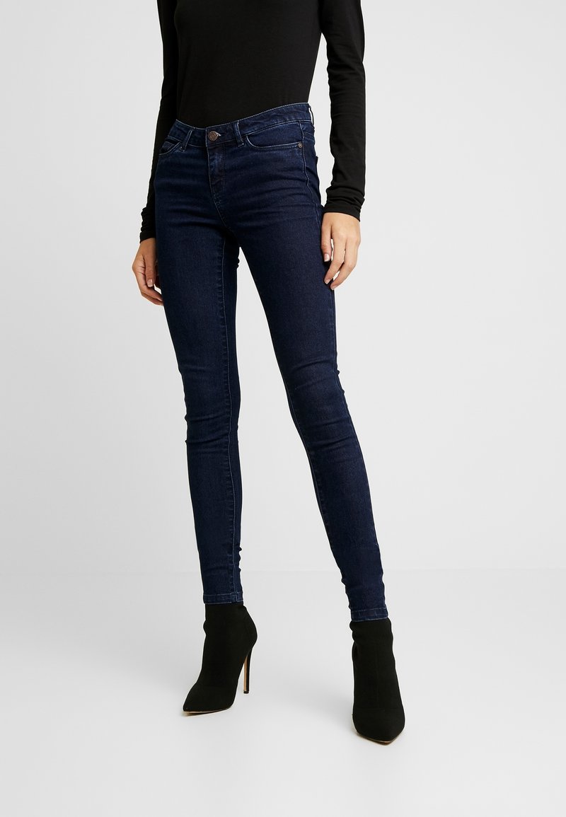 Kvinna som bär mörkblå skinny jeans, svart långärmad topp och svarta klackstövlar, står mot en enkel ljus bakgrund.