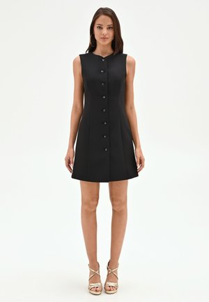 Donna in piedi che indossa un vestito nero senza maniche con bottoni e sandali dorati con tacco alto e cinturini su sfondo bianco semplice.