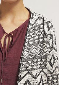 Zwart-witte open cardigan met patroon en een gestructureerd breidesign, gedragen over een bordeauxrood topje met een strikkraag.