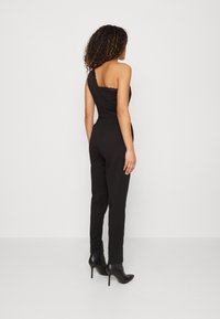Jumpsuit noir à une épaule avec un détail en dentelle au niveau du décolleté, silhouette ajustée, jambes slim, et associé à des bottines à talons hauts noires.