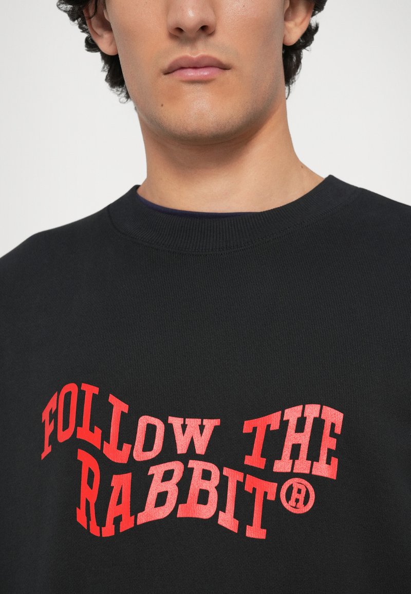 Zwarte katoenen sweatshirt met geribbelde halslijn. Heeft opvallende rode tekst met de leus "VOLG DE KONIJN" in een golvend ontwerp.