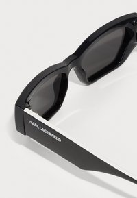 Svarte solbriller med en blank overflate, vinklet form og mørke linser. Har en hvit aksent på sideinnfatningen med "KARL LAGERFELD" gravert.
