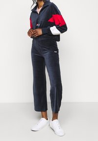 Marineblauw velours trainingspak met rood-witte accenten, cropped zip-up jas, losse broek en Fila logodetails. Gecombineerd met witte sneakers.