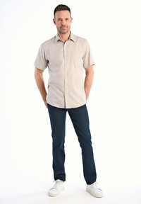 Camicia button-up beige a maniche corte con una tasca, abbinata a jeans blu scuro e sneakers bianche, in piedi davanti a uno sfondo bianco.